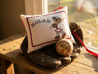 Personalized music box pillow with teddy bear design and 'Nolan' text, placed on a wooden surface with a baseball and glove.