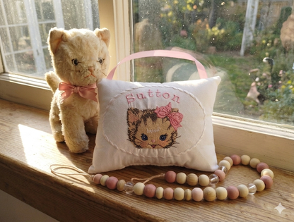 Stuffed cat and embroidered  music box pillow with a kitten design on a windowsill.