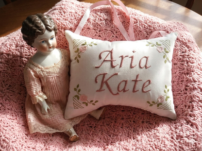 Decorative music box pillow with 'Aria Kate' embroidery next to a small doll on a pink textured surface.