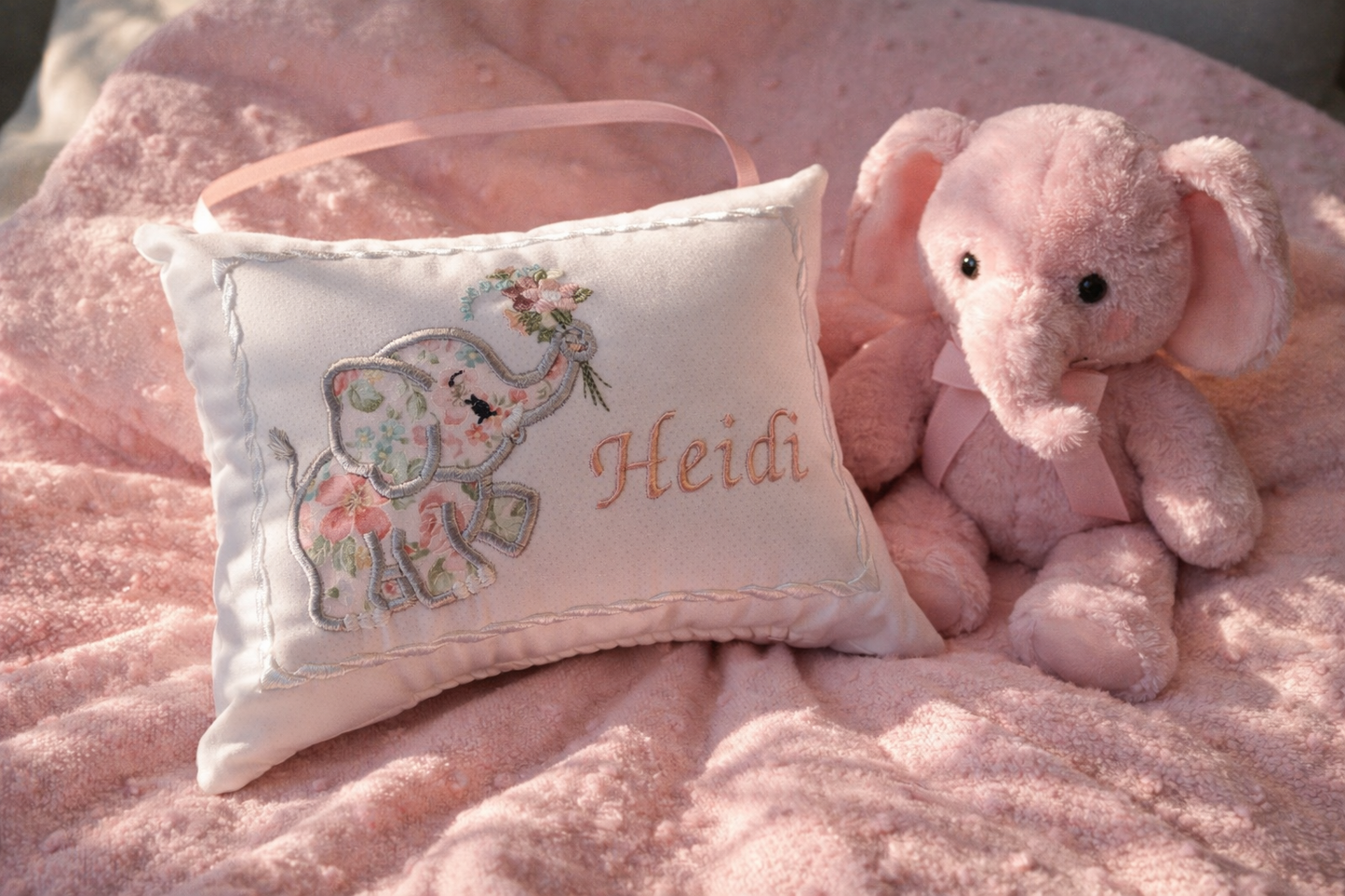 Floral elephant design music box pillow with embroidered name 'Heidi' on a pink blanket with a plush toy elephant.