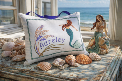 Decorative music box pillow with mermaid design and 'Aareley' embroidery, surrounded by shells and a mermaid figurine on a windowsill.