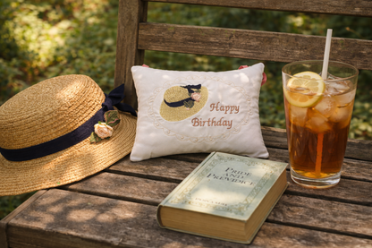 Straw hat, 'Happy Birthday' music box pillow, book, and iced tea on a wooden bench outdoors.