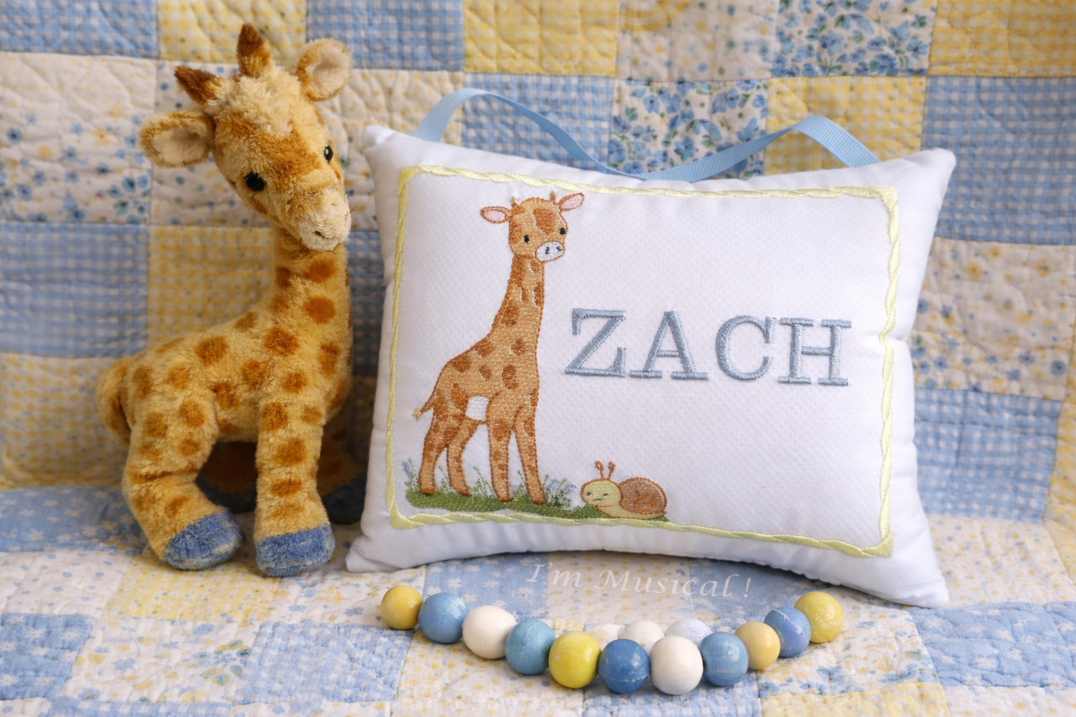 Plush giraffe toy next to a music box pillow with 'ZACH' embroidered on it, on a quilted background.