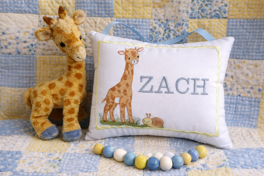 Plush giraffe toy next to a music box pillow with 'ZACH' embroidered on it, on a quilted background.