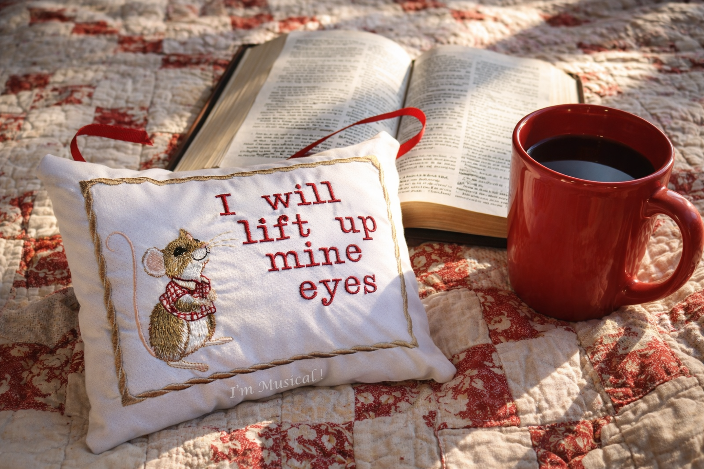 Red mug with coffee, open book, and embroidered music box mouse pillow on a quilt.