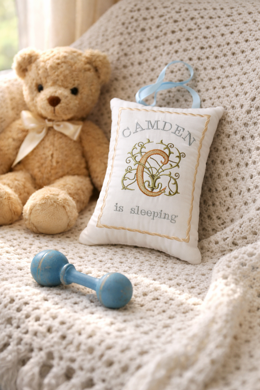 Baby music box pillow with 'Camden is sleeping' text, teddy bear, and blue rattle on a textured surface.