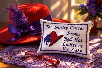 Red hat with purple ribbon and feather next to a embroidered music box  pillow with text, on a doily.