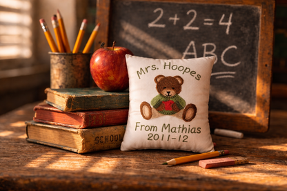 Decorative music box pillow with teddy bear and text, books, apple, and chalkboard in the background