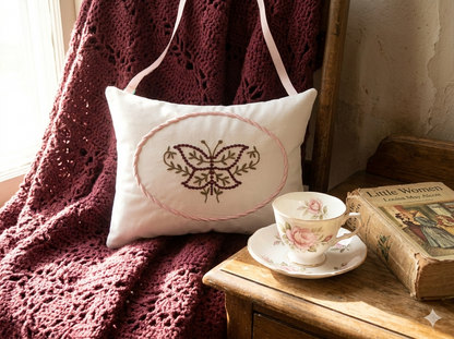 Decorative music box pillow with embroidered butterfly design on a wooden surface with a teacup and books.