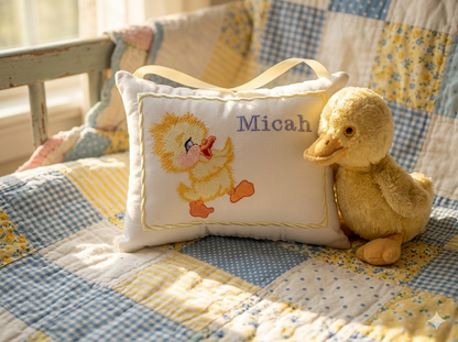 Decorative music box pillow with a duck design and embroidered name 'Micah' on a patchwork quilt, accompanied by a plush duck toy.
