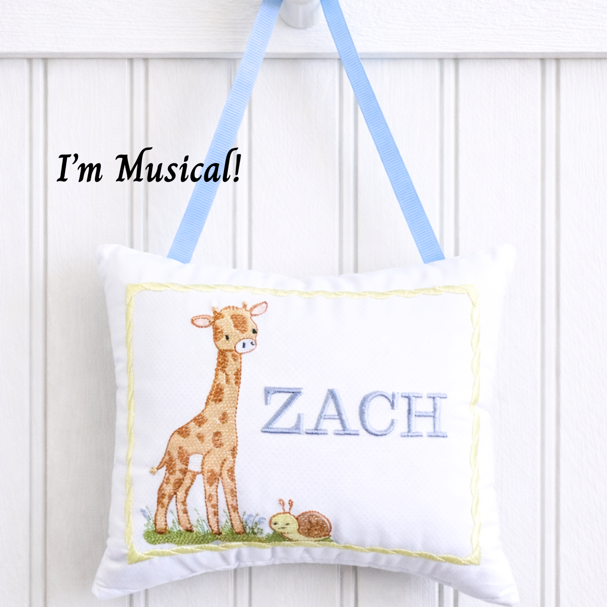 Children's music box pillow with an embroidered giraffe design and the name 'Zach' on a white background.