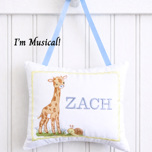 Children's music box pillow with an embroidered giraffe design and the name 'Zach' on a white background.