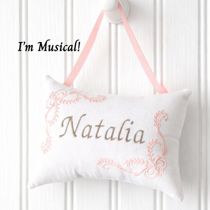 Decorative music box pillow with embroidered name 'Natalia' and pink ribbon on a white background