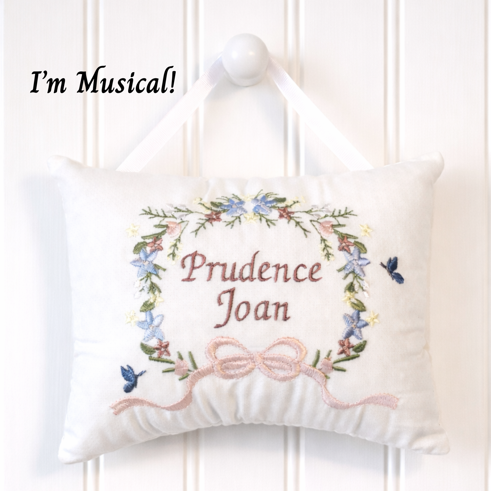 Decorative music box pillow with floral design and embroidered text 'Prudence Joan' on a white background.