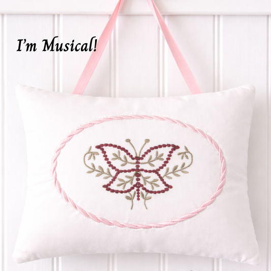 White music box pillow with embroidered butterfly design and pink straps on a white background