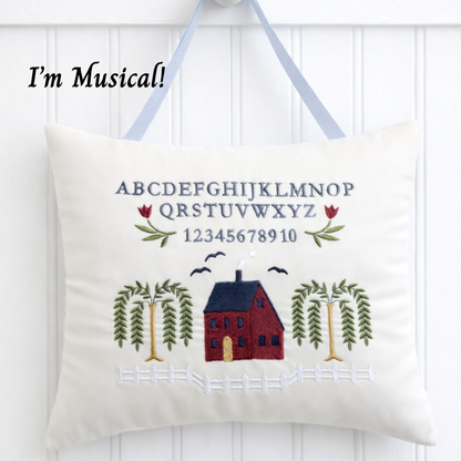 Decorative music box pillow with embroidered alphabet, numbers, house, trees, and birds on a white background.