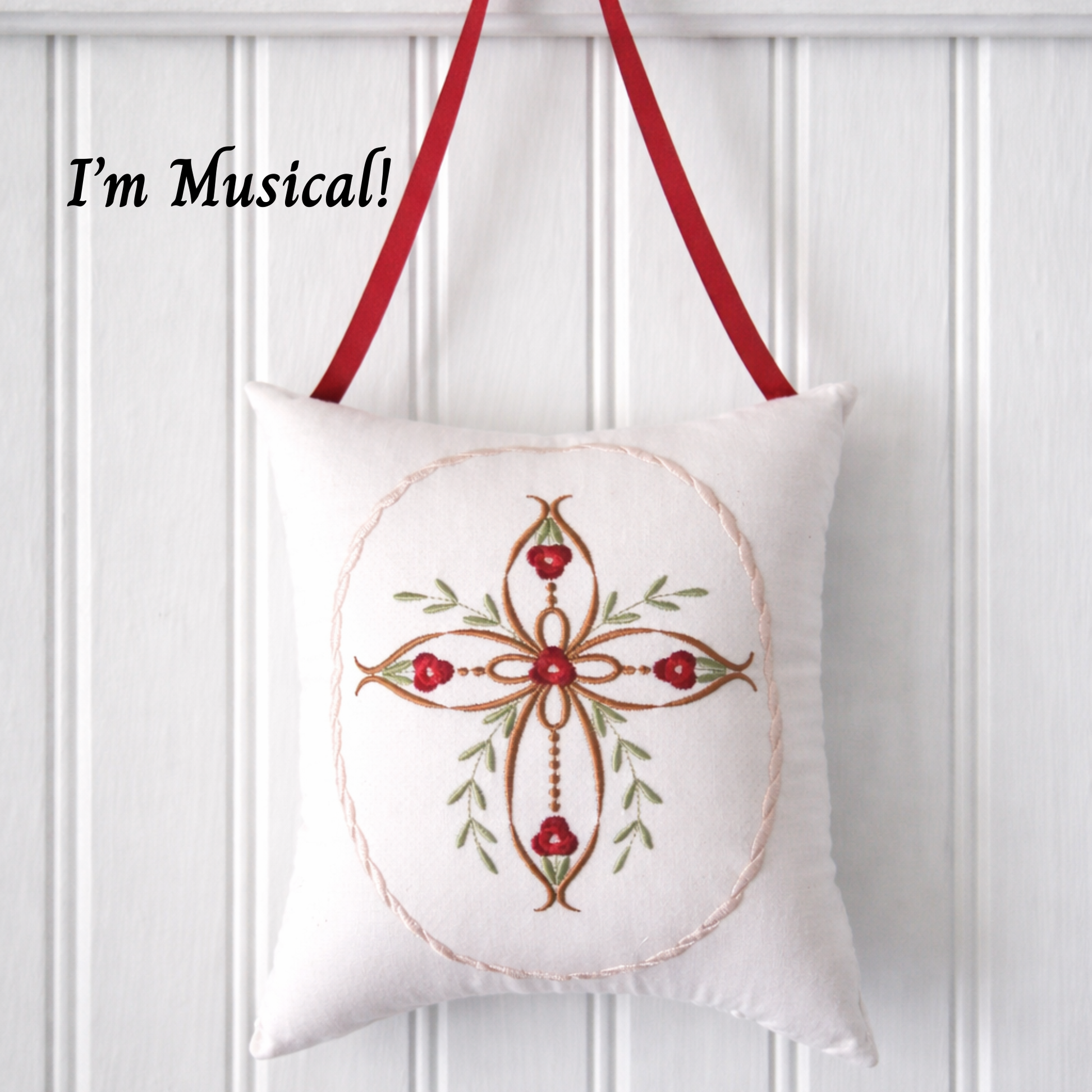 Decorative music box pillow with embroid cross and red roses on a white paneled wall.