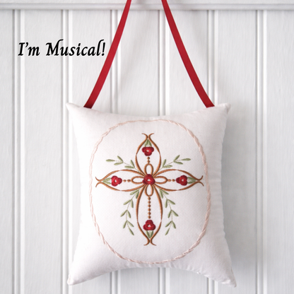 Decorative music box pillow with embroid cross and red roses on a white paneled wall.