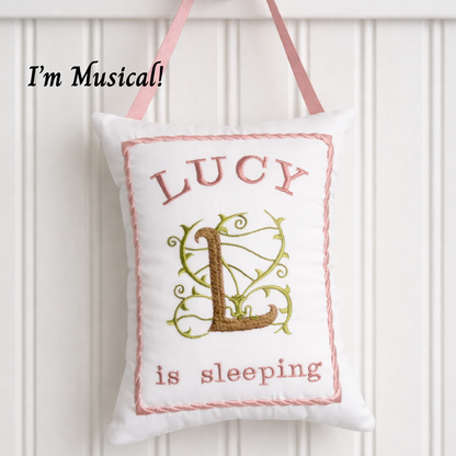 Decorative music box pillow with 'LUCY is sleeping' text and a decorative letter 'L' on a white background.