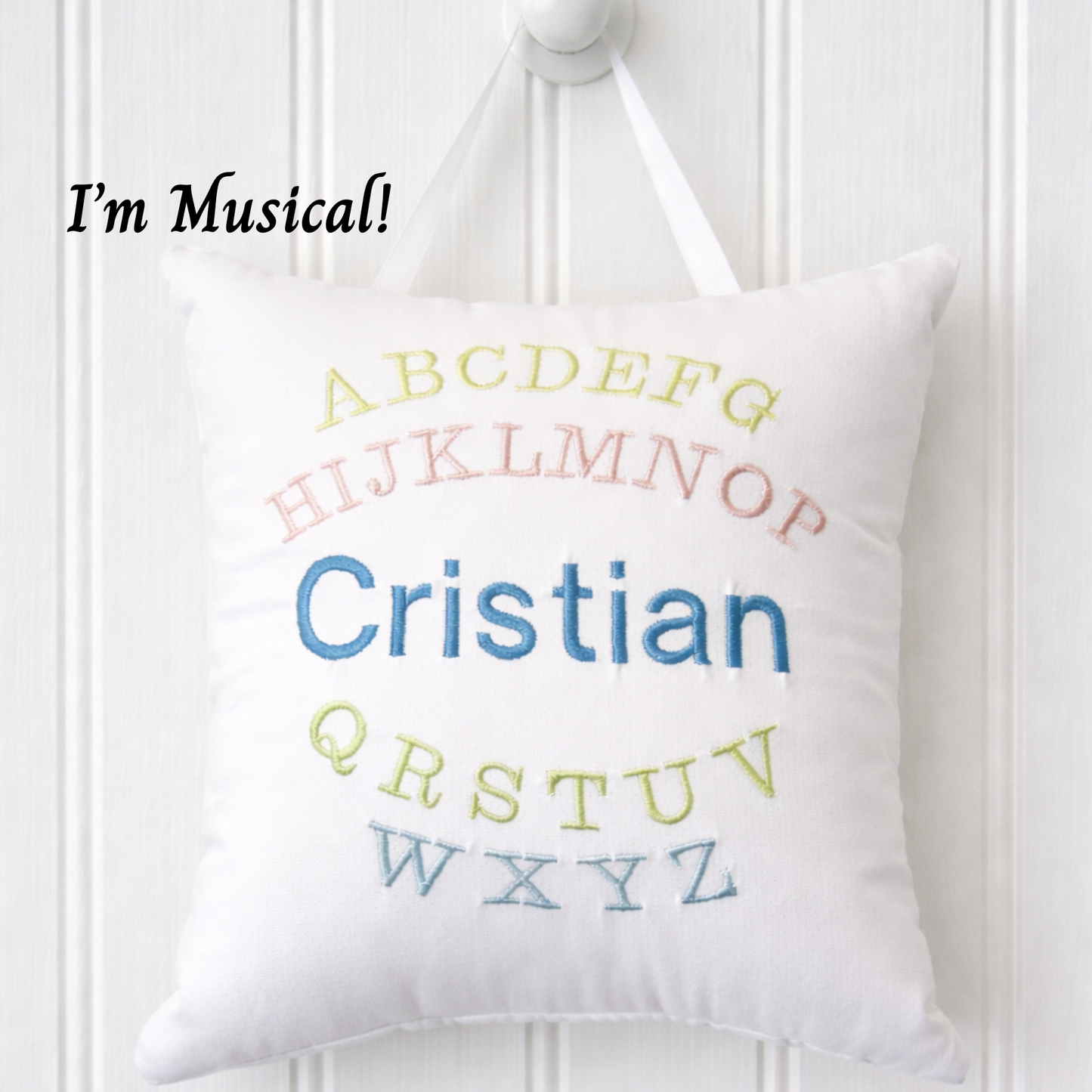 White music box pillow with embroidered colorful letters and name 'Cristian' on a white wooden background.