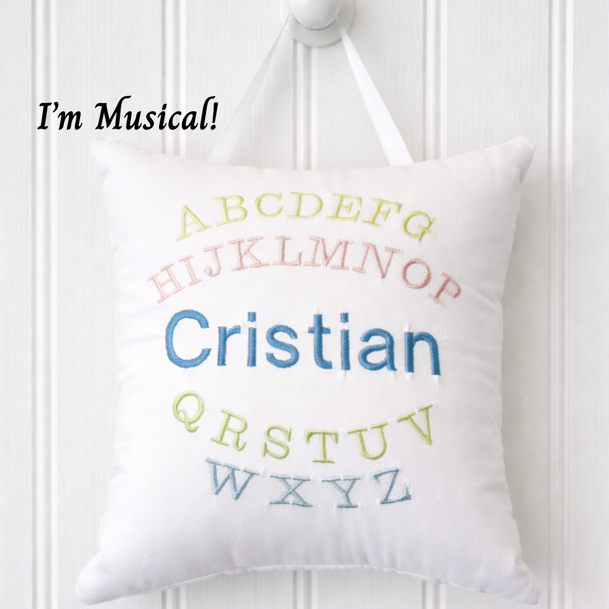 White music box pillow with embroidered colorful letters and name 'Cristian' on a white wooden background.