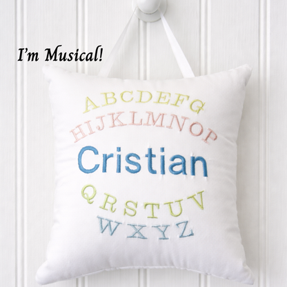 White music box pillow with embroidered colorful letters and name 'Cristian' on a white wooden background.
