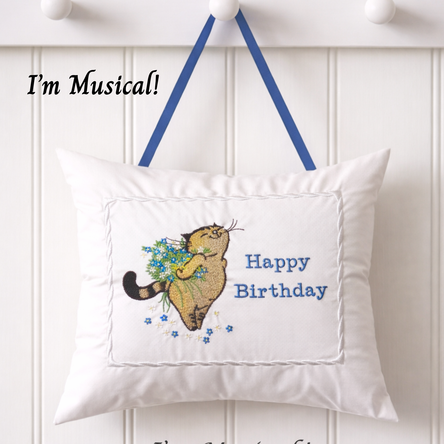Decorative music box pillow with a cat holding flowers and 'Happy Birthday' text, hanging on a white wall.