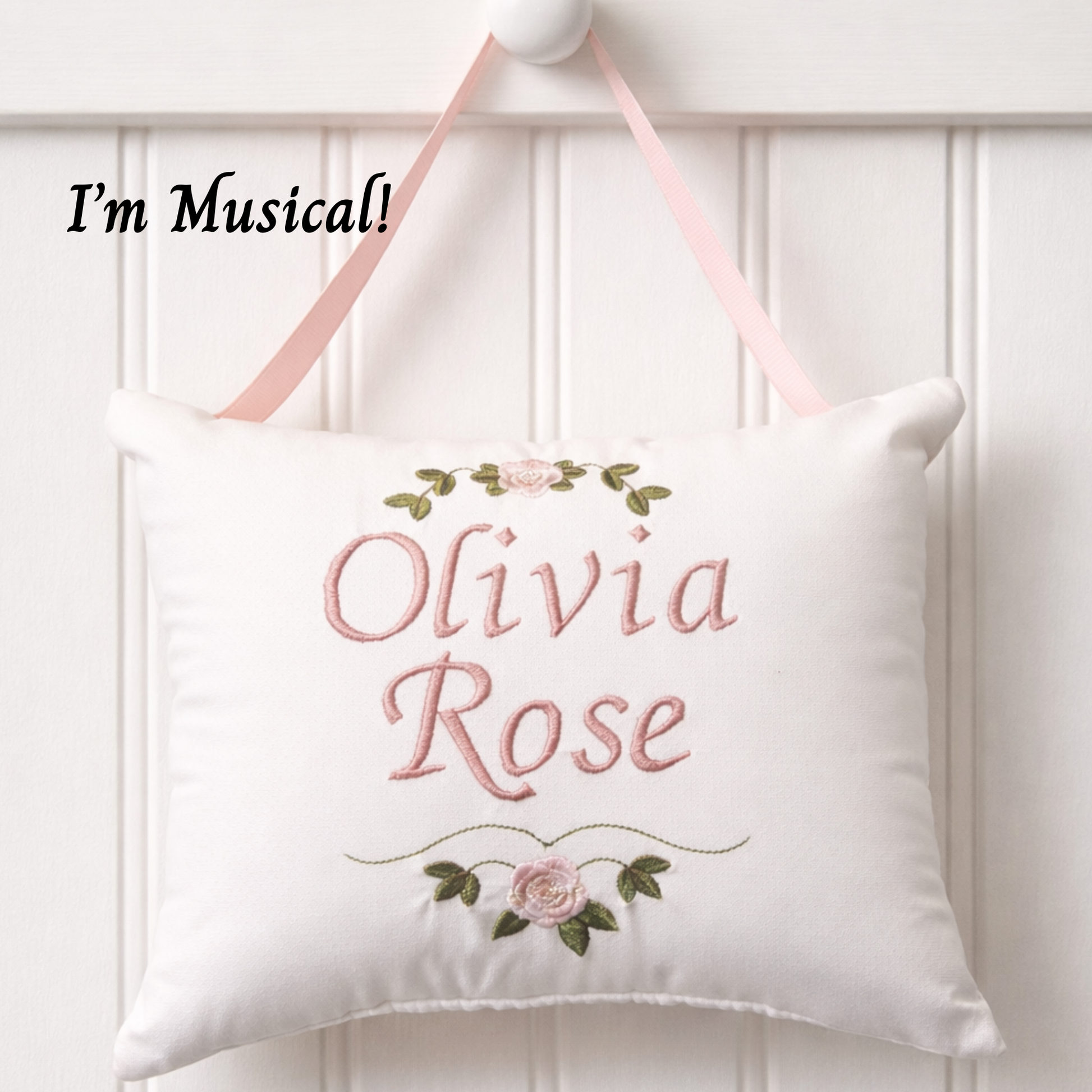Decorative music box pillow with embroidered name 'Olivia Rose' and floral designs, hanging against a white wooden panel background.