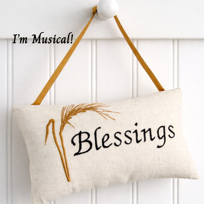 Decorative music box pillow with 'Blessings' text and straw design, hanging against a white wooden panel background.