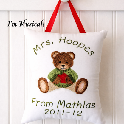 Decorative music box pillow with embroidered teddy bear and text, hanging against a white background.