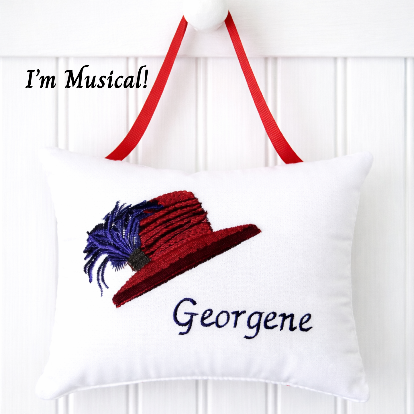 White music box pillow with a red and blue hat design and 'Georgene' text, hanging against a white background.