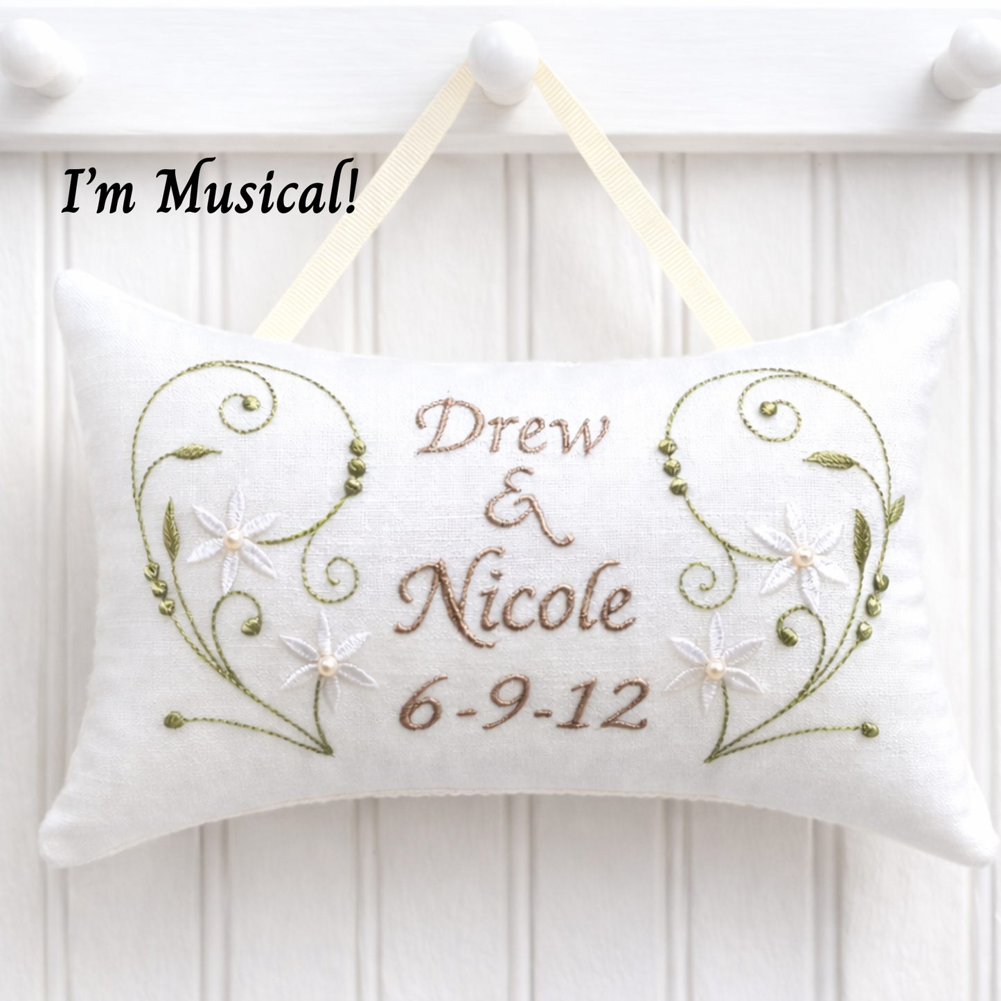 Decorative music box pillow with embroidered text 'Drew & Nicole 6-9-12' on a white wooden background.