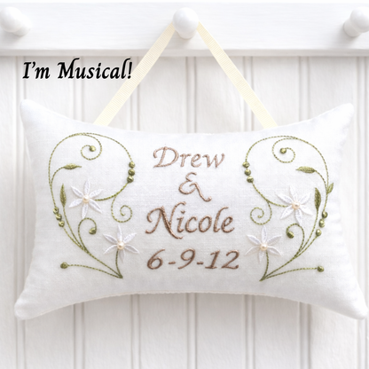 Decorative music box pillow with embroidered text 'Drew & Nicole 6-9-12' on a white wooden background.