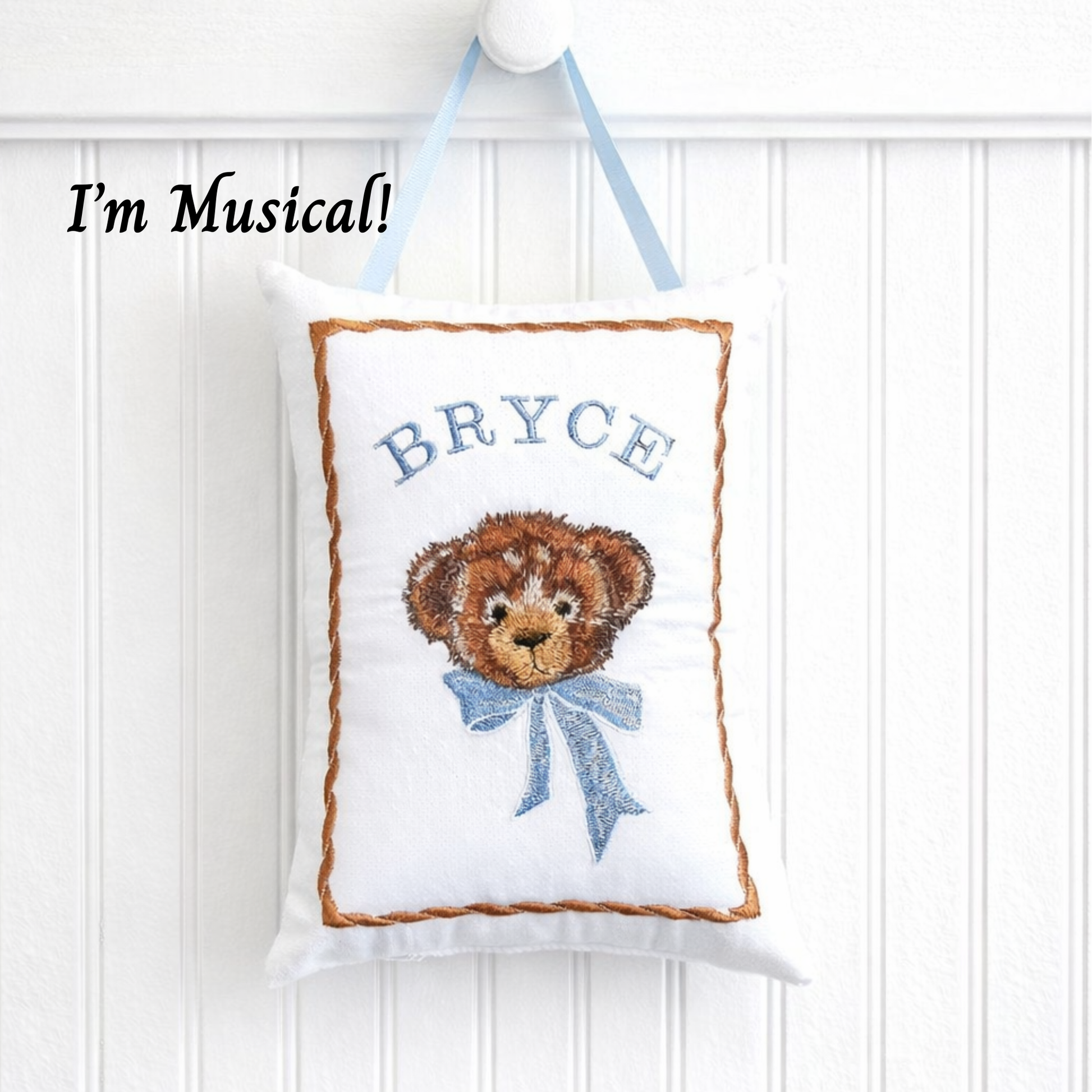 Decorative music box pillow with teddy bear design and 'BRYCE' text, hanging against a white wall.