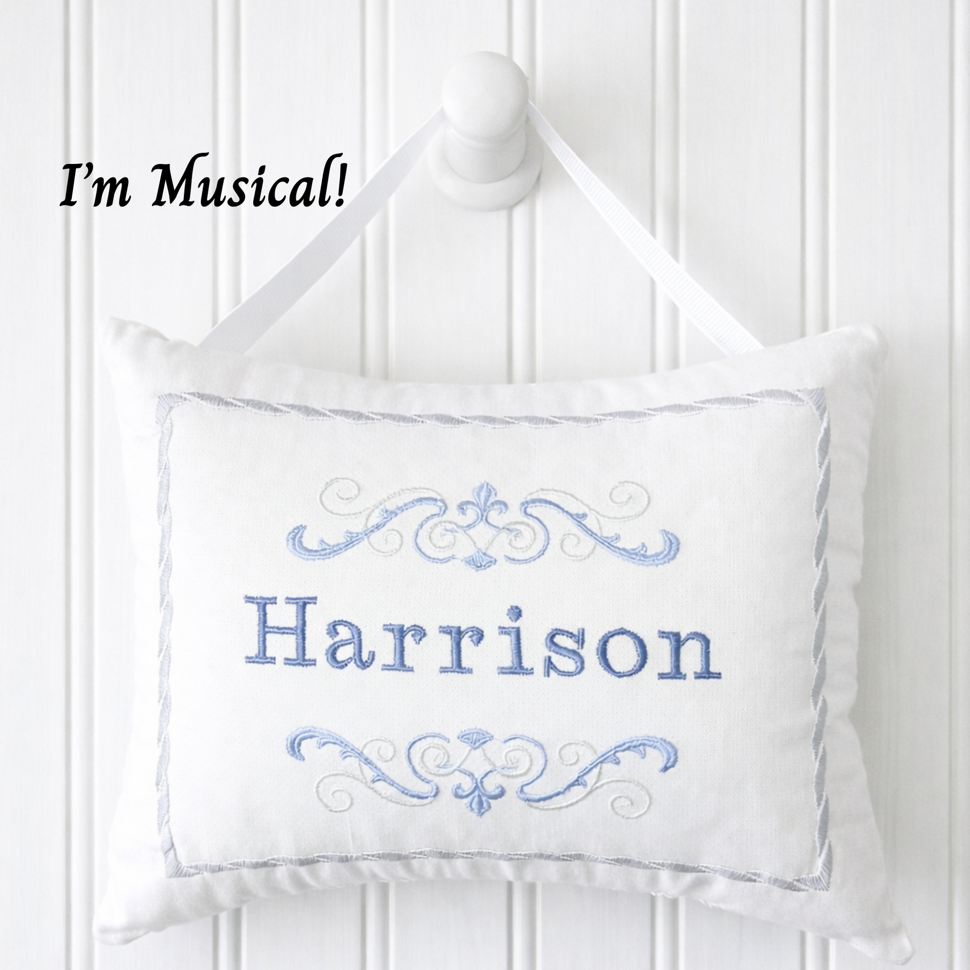 White music box pillow with blue embroidery and 'Harrison' text on a white wooden panel background