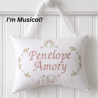 Decorative music box pillow with embroidered names 'Penelope' and 'Amory' on a white wooden background.