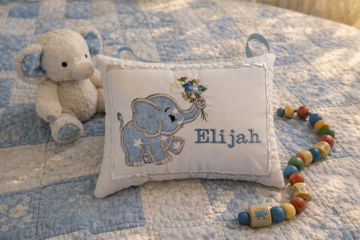 Decorative music box pillow with an elephant design and embroidered name 'Elijah' on a quilted blanket with a plush elephant and beads.