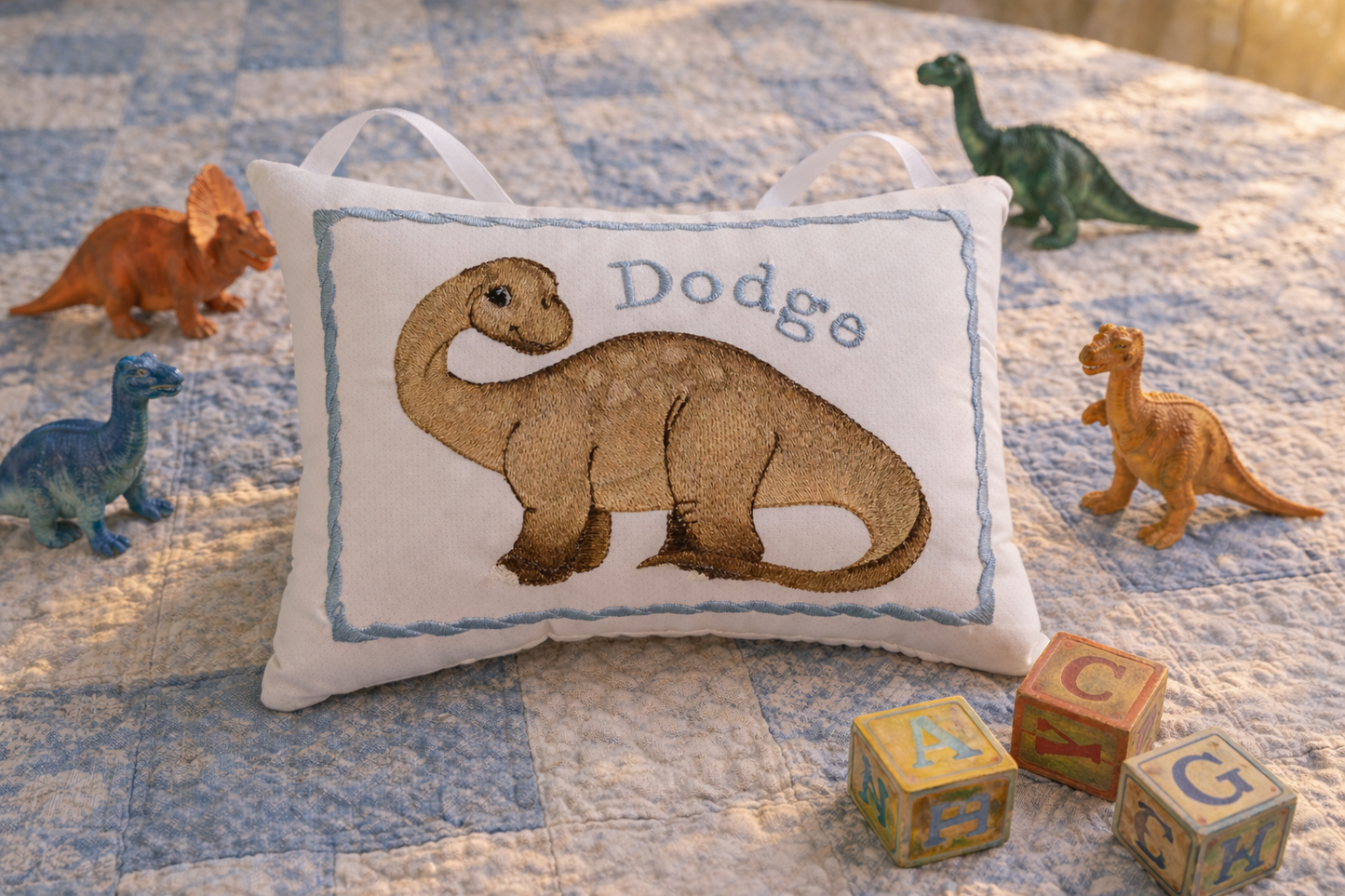 Decorative music box pillow with a dinosaur design and embroidered name on a bed with toy dinosaurs and blocks.