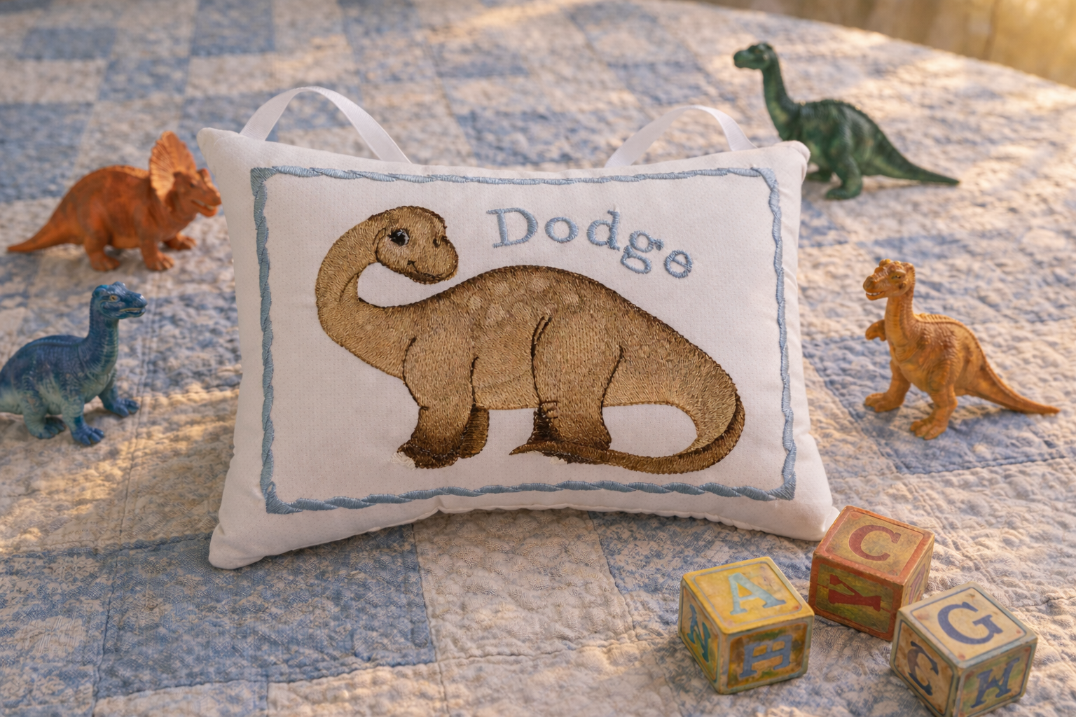 Decorative music box pillow with a dinosaur design and embroidered name on a bed with toy dinosaurs and blocks.