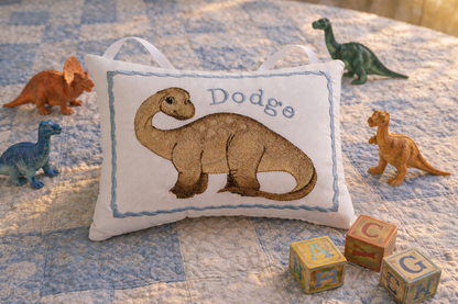 Decorative music box pillow with a dinosaur design and embroidered name on a bed with toy dinosaurs and blocks.
